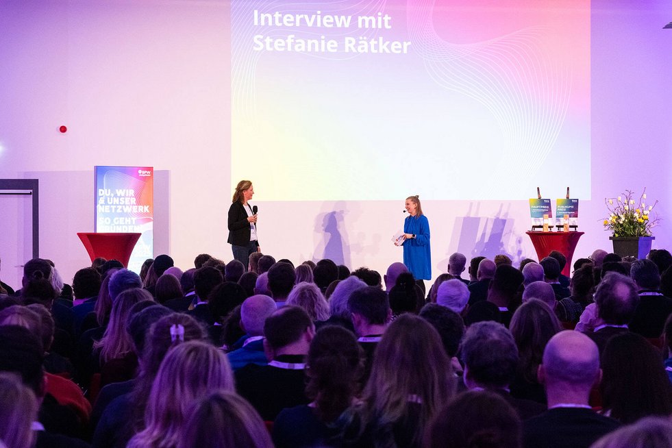 zwei Personen sprechen auf einer Bühne miteinander, ein Publikum hört in einem Saal zu