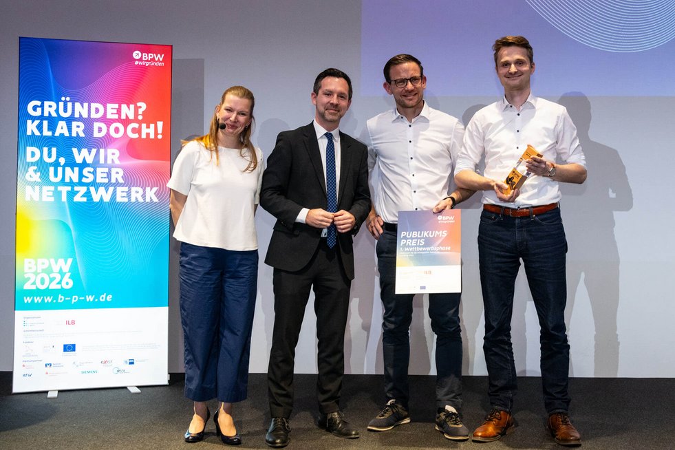 Vier Personen stehen auf einer Bühne bei einer Preisverleihung. Zwei Personen halten eine Urkunde und eine Trophäe. Links ist ein Roll-up mit der Aufschrift „Gründen? Klar doch! BPW 2026“ zu sehen.