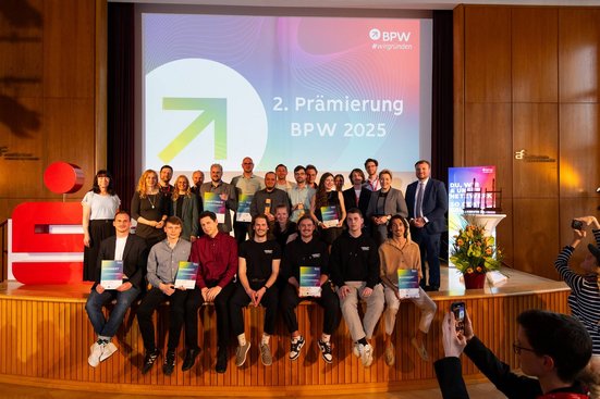 Gruppenfoto mit vielen Menschen, die entweder stehend oder auf einer Bühne sitzend Richtung Fotograf schauen