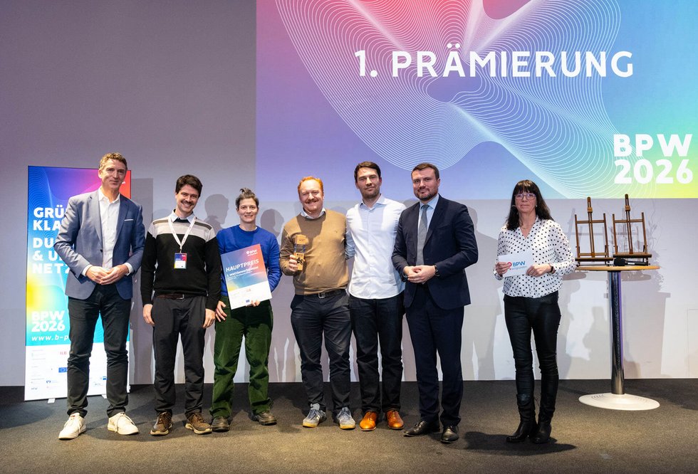 Gruppe von acht Personen auf einer Bühne bei einer Preisverleihung. In der Mitte halten zwei Personen eine Urkunde und eine Trophäe. Im Hintergrund ist eine Projektion mit der Aufschrift „1. Prämierung – BPW 2026“ zu sehen.